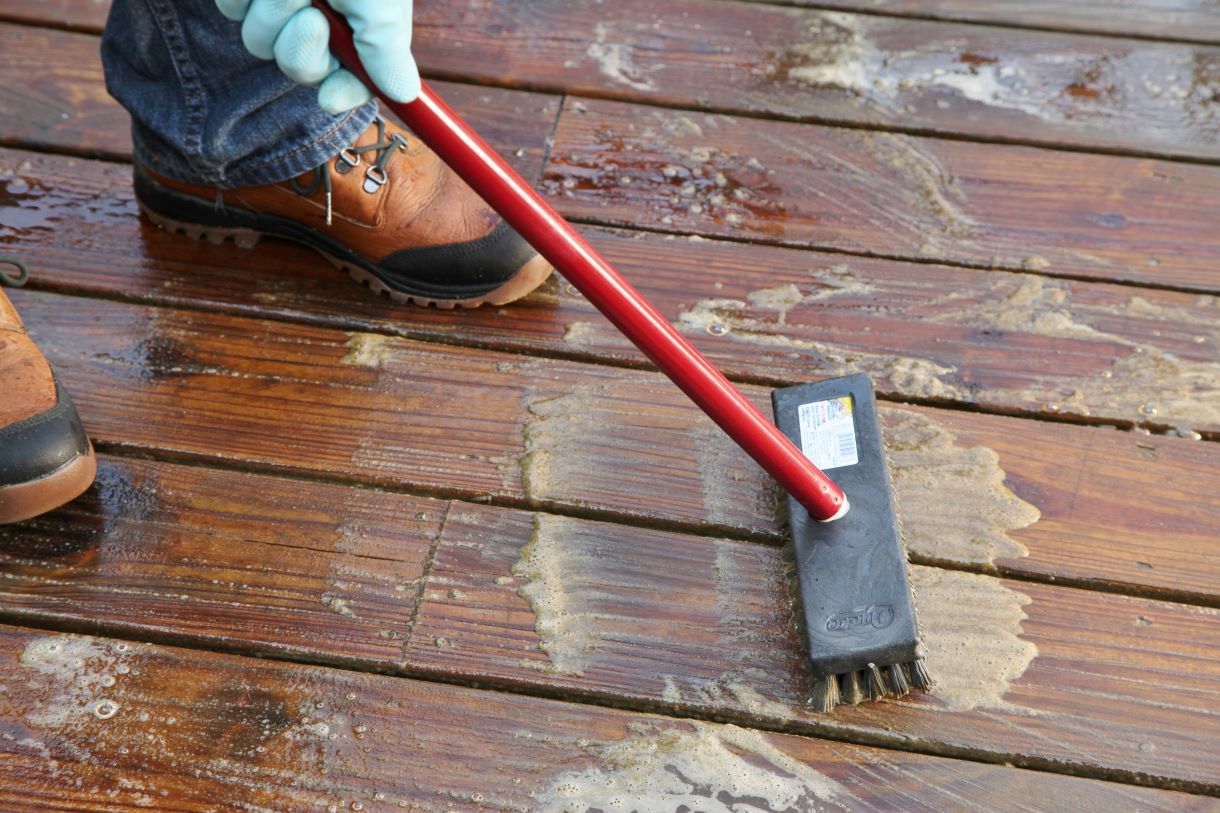 Close-up of a person using a stiff-bristle brush to clean a wooden deck with Thompson's WaterSeal Heavy Duty Deck Cleaner, preparing the surface for waterproofing.